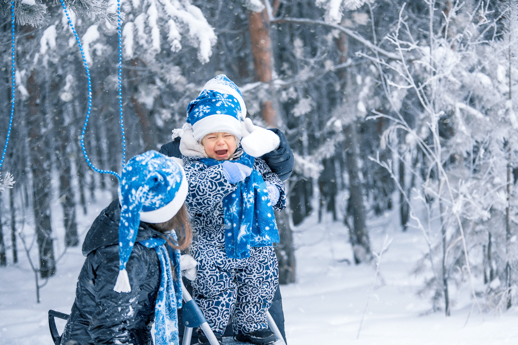 A baby in a Santa Claus hat is crying surrounded by his sisters against the background of a snowy forest. Winter time in the forest.